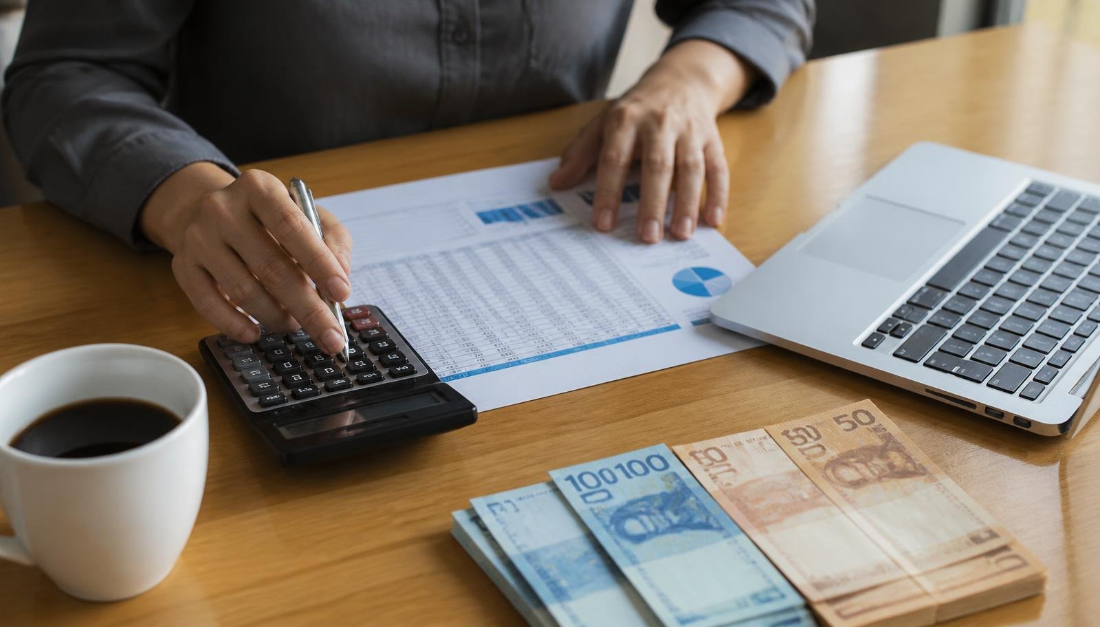 Pessoa fazendo contas com calculadora, planilha impressa, notebook e notas de real sobre a mesa durante planejamento de investimentos